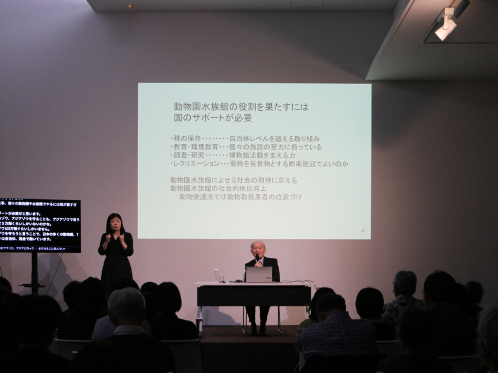 講演会で中央に座る成島氏、左手に立つ手話通訳者